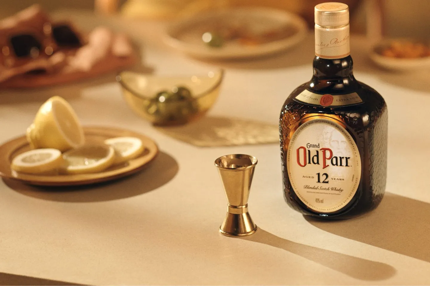Garrafa de Old Parr 12 anos sobre a mesa, ao lado de um jigger e de pratos com limão e azeitona