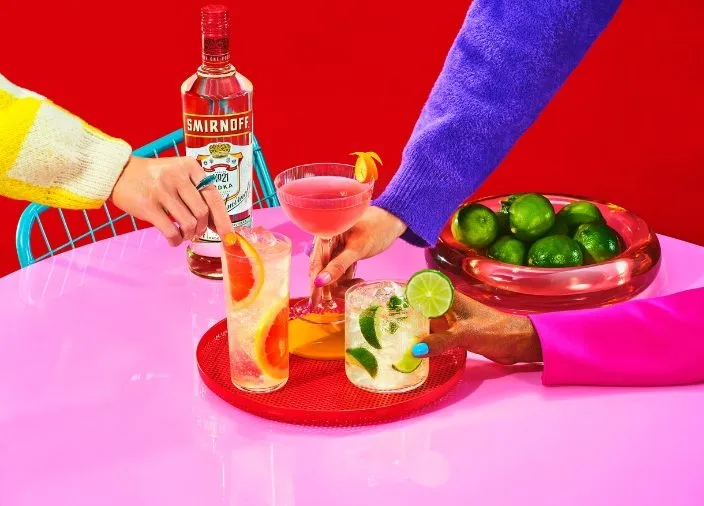 Mãos pegando três coquetéis coloridos numa mesa, com a garrafa de Smirnoff ao fundo