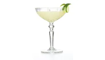 Coquetel de cor amarelo-pálido servido em uma taça coupe de cristal lapidado com haste elegante. Uma casca de limão verde em espiral decora a borda direita. O fundo é totalmente branco, isolando a bebida.