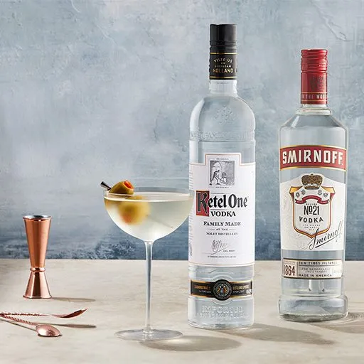 Uma garrafa de vodka Ketel One e vodka Smirnoff em uma mesa, ao lado de um copo de coquetel clássico, contendo uma bebida gelada e uma guarnição de duas azeitonas. Um dosador e um mixer em ouro rosa estão sobre a mesa.
