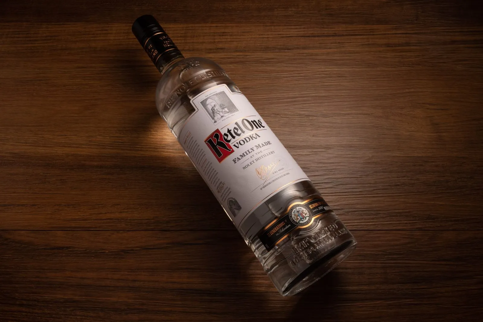 Garrafa de vodka Ketel One deitada na diagonal sobre superfície de madeira escura. A iluminação destaca o rótulo branco com a marca em vermelho e preto, além dos detalhes em relevo no vidro. Textura de madeira rica e sombras suaves compõem o cenário.