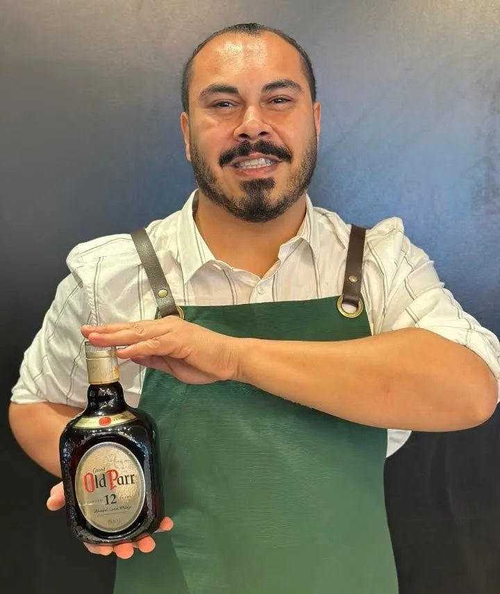 Henrique Abrahão de avental verde segurando uma garrafa de Old Parr 12 anos