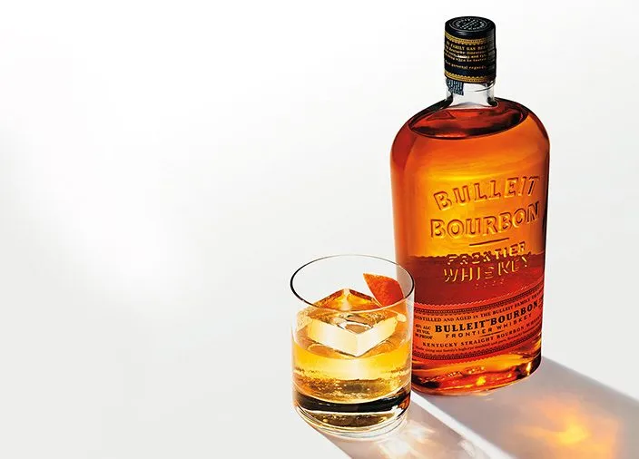 Uma garrafa de Bulleit Bourbon em uma mesa branca ao lado de um copo cheio de Bulleit e um grande cubo de gelo. Sombras brilhantes são projetadas no canto inferior direito. 