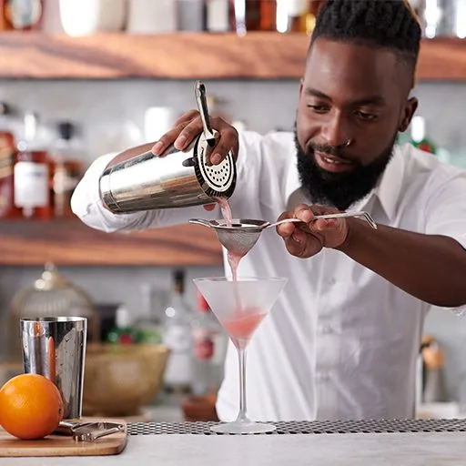 Bartender despejando um coquetel rosa através de um coador em uma taça de Martini gelado. 